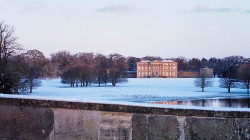 A large historic mansion sits in the distance, surrounded by a snow-covered landscape and bare winter trees. The building has a symmetrical design with tall windows and a classical facade. In the foreground, a stone wall runs horizontally across the image, and a reflective body of water lies just beyond it. The sky above is pale with soft pink and blue hues, suggesting early morning or late afternoon light.